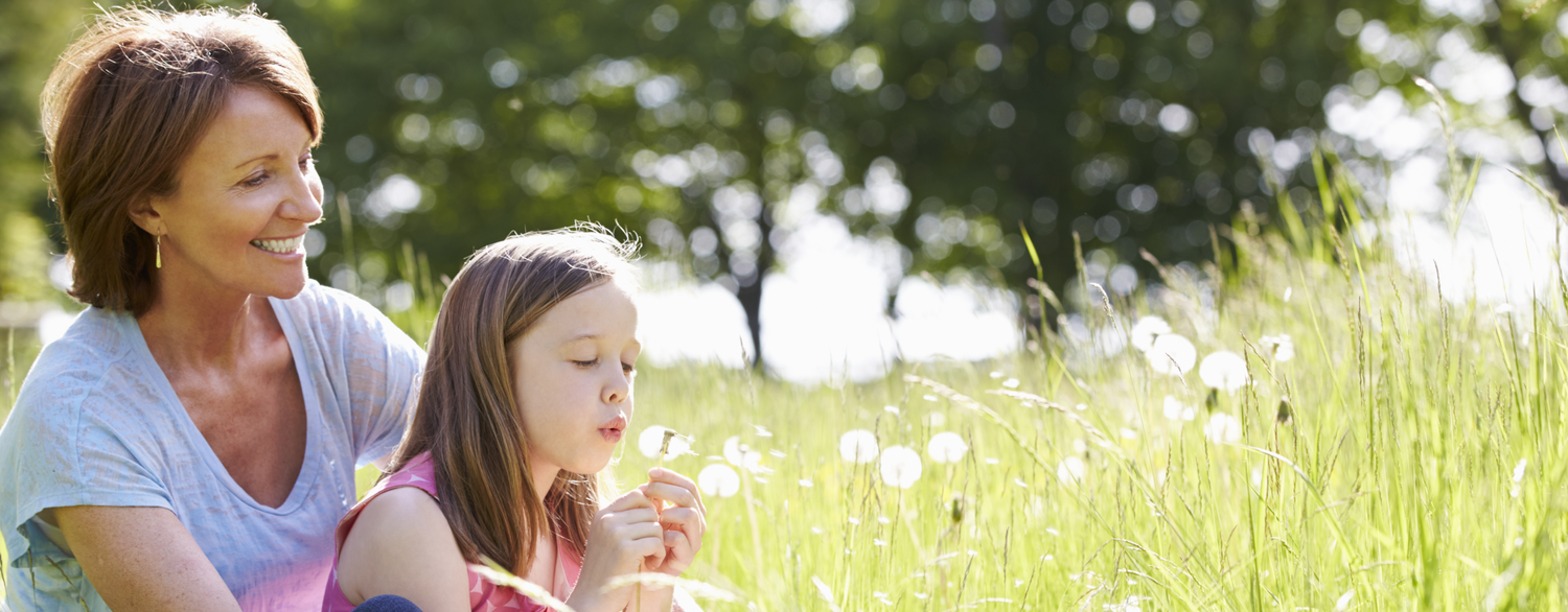 A woman sitting in a field with a young girl who is blowing fuzz off a dandelion.